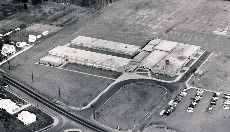 Black and white aerial photograph of Mark Twain Intermediate School taken during the 1960s. The building has two, long rectangular classroom wings, extending outward, and parallel to one another, from a square-shaped section of the building which houses the cafeteria, offices, and gymnasium. 