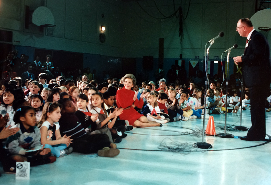Photograph of the school assembly during Nancy Reagan’s visit to Cameron. She is seated on the floor along with the students. Principal Towery is standing at a microphone addressing those in attendance.