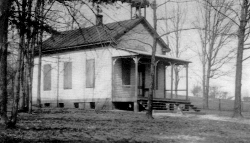 Black and white photograph of the Cameron School, a one-room schoolhouse with white siding and a pitched tin roof. A wooden porch covers the main entrance. The building has three windows on each side, and the shutters are closed. There is a bell tower and brick chimney on the roof of the structure. The school is set on a hillside and is surrounded by trees. A small farmhouse is visible in the distance. 