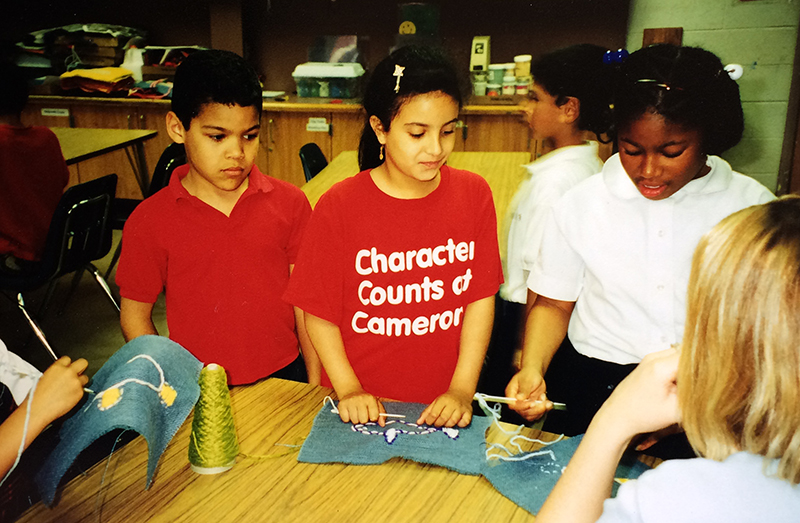 Photograph of three students participating in a classroom activity. They are wearing school uniforms. The child in the center is wearing a Character Counts at Cameron t-shirt.