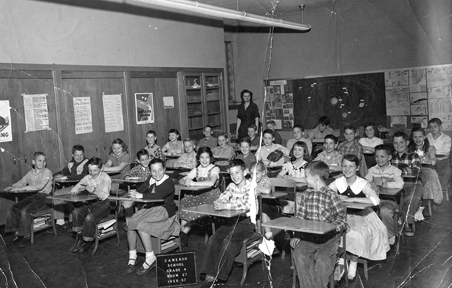 Black and white class portrait of a fourth grade room during the 1956 to 1957 school year. A sign on the floor indicates this is Classroom 27. 30 children are pictured, seated at their desks. A female teacher is in the far back corner of the room.
