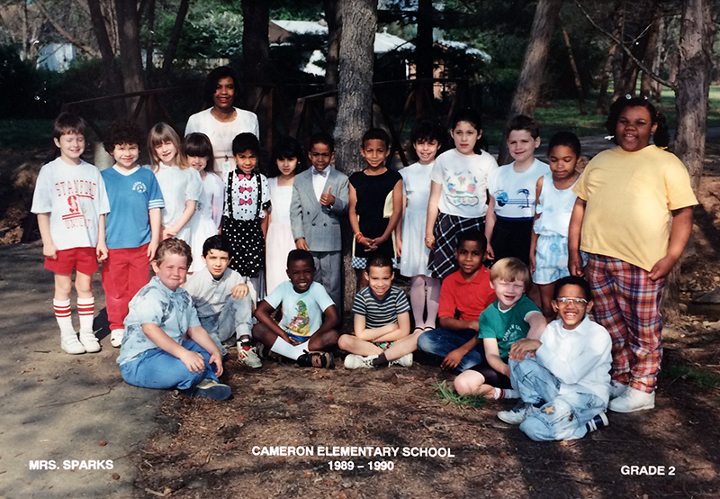 Cameron Elementary School class photograph showing Mrs. Spark’s class of second graders. 20 students and their teacher are pictured. They are standing on a path in a forest, probably on the school camping trip.
