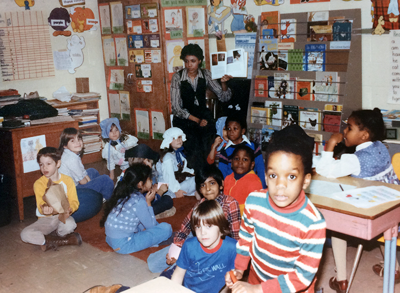 Color photograph of a Cameron Elementary School classroom. Based on the clothing, the picture was probably taken during the early 1980s. The children are sitting on the floor in front of a teacher who is holding up a book.