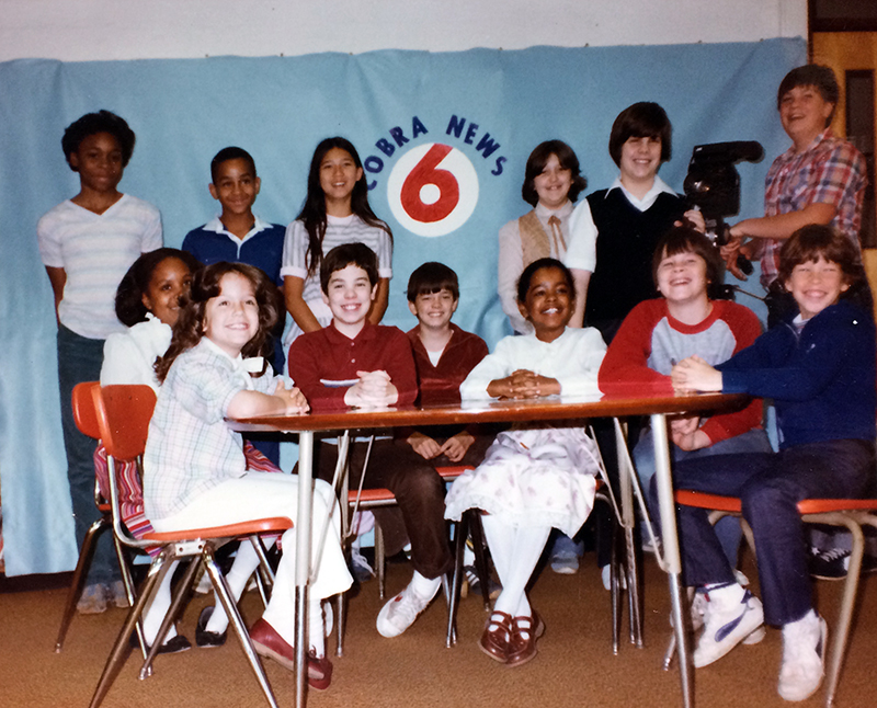 Color photograph of the Cameron Cobra News Team taken in the late 1980s or early 1990s. 13 children are pictured. They are posed on the news set and one of them is operating a video camera.