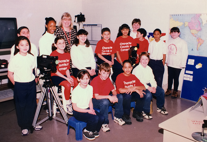 Color photograph of the Cameron Cobra News Team taken in the late 1990s or early 2000s. The technology in the news room includes a television, VHS camcorder, tripod, and microphones. There is a world map on the wall. 15 children, wearing school uniforms, and a teacher are pictured.