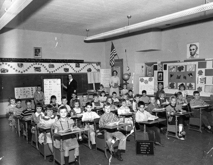 Black and white class portrait of a first grade room during the 1956 to 1957 school year. A sign on the floor indicates this is Classroom 4. 33 children and two female adults are pictured. Most of the children are seated at their desks, but five children at the back of the room are standing. Both of the women are at the back of the room. One is standing next to the chalkboard and the other is standing next to a doorway.