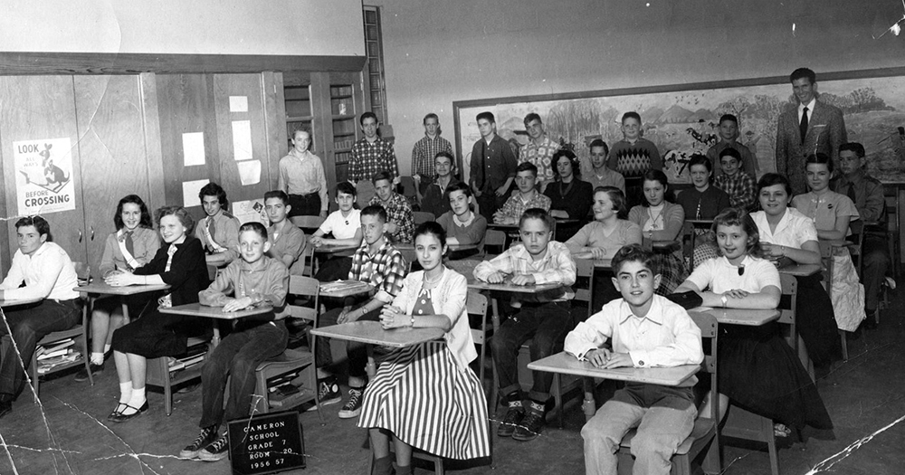 Black and white class portrait of a seventh grade room during the 1956 to 1957 school year. A sign on the floor indicates this is Classroom 20. 32 children and one male adult are pictured. Most of the children are seated at their desks, but seven boys are standing along the wall at the back of the classroom. The teacher is standing in the back on the far right side of the picture.