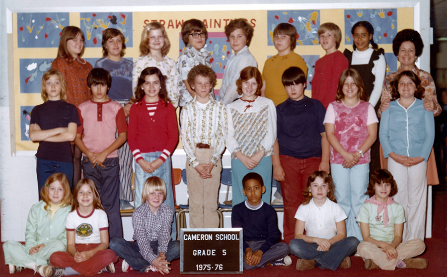 Color class portrait of a fifth grade room during the 1975 to 1976 school year. 22 children and one female teacher are pictured. The children are arranged in three rows. The first row is seated on a red carpet floor, the second row is standing, and the third row is standing on chairs. The teacher is in the third row, on the far right side of the image. They are posed in front of a wall inside the school. A hallway billboard featuring student artwork is behind them.