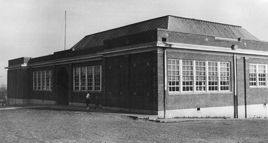 Black and white photograph of Lee-Jackson High School. The school is a two-story brick building built into a hillside so that from the front view of the main entrance the building appears only one-story tall. Large banks of windows line three sides of the building. According to FCPS records, the windows were a frequent target of rock-throwing vandals. There is a raised section at the center of the building with a pitched roof, indicating there may have been an auditorium is this area. The main entrance doors are reached through a tall, brick archway. In this view of the front of the building, taken in 1942, two people can be seen standing under the windows at the front of the building.