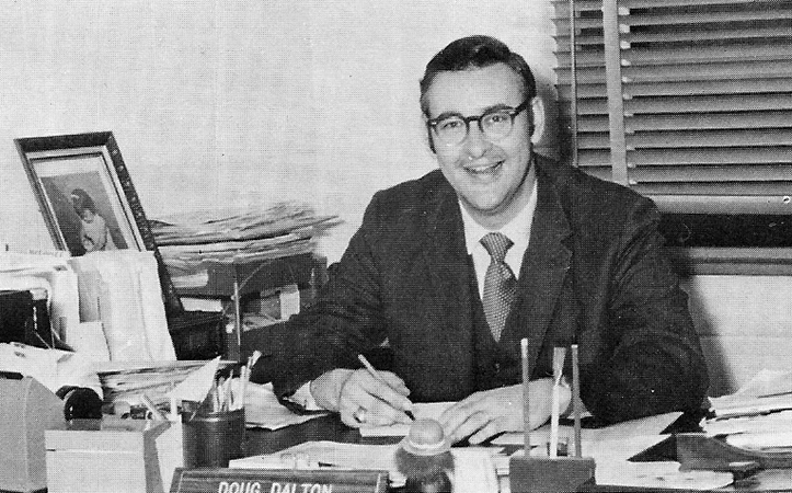 Black and white yearbook photograph of Principal Dalton taken during the 1972 to 1973 school year. He is seated at his desk and is looking up from paperwork that he is in the process of signing.
