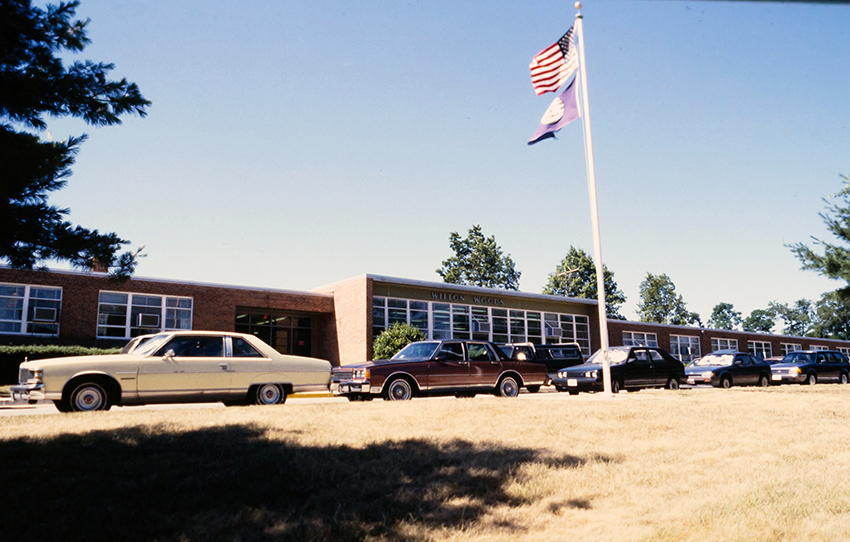 Color photograph of the front exterior of Wilton Woods Elementary School from a 35 millimeter slide. The picture appears to date from the late 1970s or early 1980s. Wilton Woods is a single-story brick structure with banks of windows facing along Franconia Road. Window-mounted air conditioning unit are in place in every room. Several cars are parked along the driveway in front of the building.